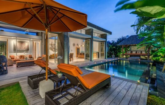 Swimming pool view with day beds and villa exterior with lights on in background