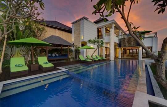 Large swimming pool of villa at dusk with villa exterior and day beds