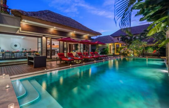 View of swimming pool at dusk with lights on with villa exterior in background