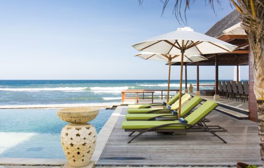 Day beds by the pool with umbrellas and the ocean in the background