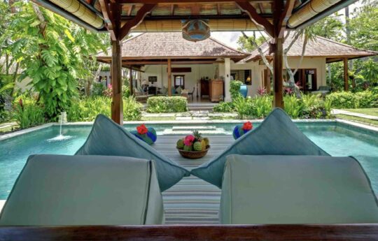 View of swimming pool from day bed area on a sunny day, villa exterior in background