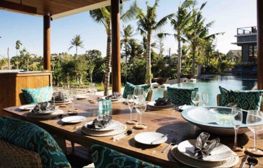 Dining table of villa with seating for 8 people with view of pool and garden on sunny day